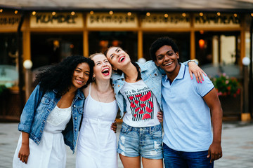 Group of four multiethnic hipster teenagers friends laughing and hugging while spending time together outdoor in ciity center
