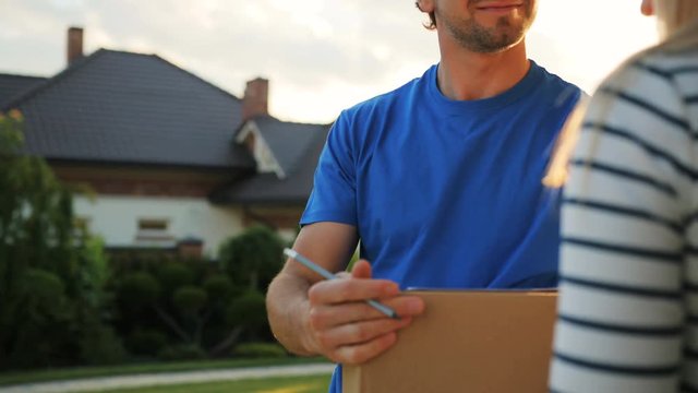 Young courier delivering the cardboard to the house, happy woman receiving the package and signing the documents for getting this. Close up shot.