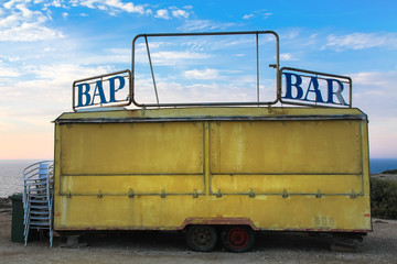 Old Yellow Bar Trailer vehicle with word Bar partially broken on the left side of the frame against a blue background sky with white clouds during sunset.