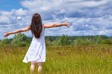 Naklejka na ściany i meble Girl in white dress standing in the field enjoying privacy. Bright blue clouds in the sky, the green grass with spikes
