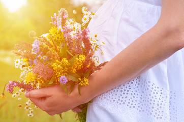 Pregnant young girl stands in the field hugging her stomach by hand with a bouquet of wildflowers. The concept of motherhood and caring, sunlight