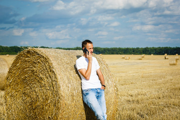 Naklejka na ściany i meble Handsome young man at countryside, talking on cell phone, standing in field, wearing white T-shirt and jeans,