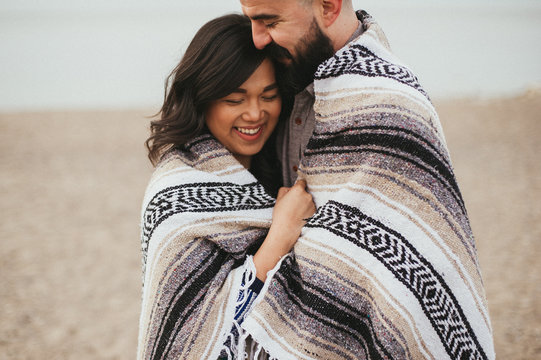 Couple wrapped in Mexican blanket and hugging and smiling on beach