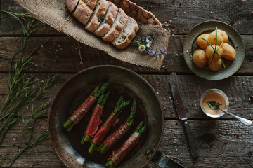 green asparagus wrapped in prosciutto ham, potatoes, hollandaise sauce and olive bread on wooden background
