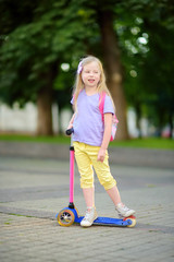 Little child learning to ride a scooter in a city park on sunny summer day. Cute preschooler girl...