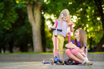 Small children learning to ride scooters in a city park on sunny summer evening. Cute little girls...