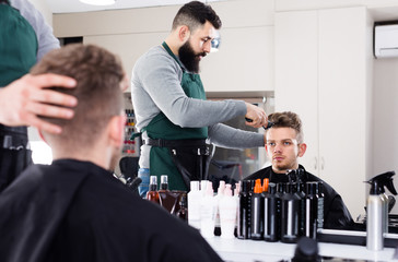 Young male stylist creating haircut for man client at hairdressing salon