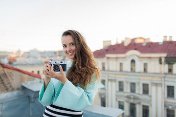 Fashion look, pretty cool young woman model with retro film camera. curly hair outdoors. Stylish girl photographer takes the old city from the roof at sunset.