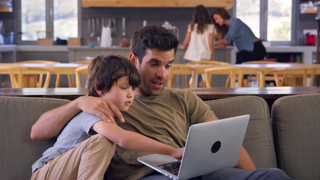 Father And Son Sitting On Lounge Sofa Using Laptop Together