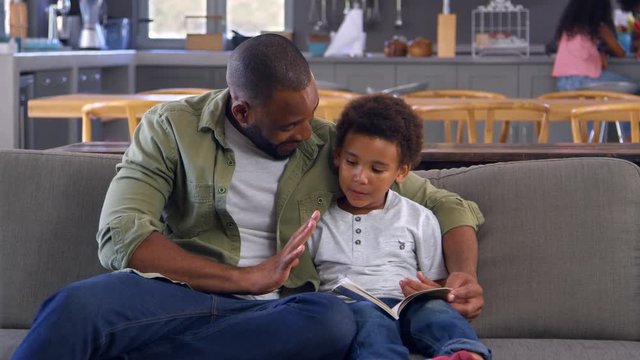 Father And Son Sitting On Sofa Looking At Counting Book Together