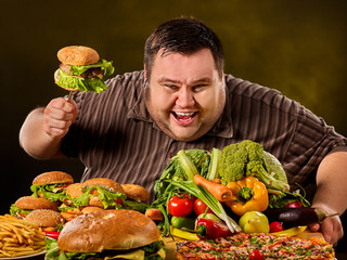 Diet fat man who makes choice between healthy and unhealthy food. Overweight male with hamburgers. Table with food in the foreground.