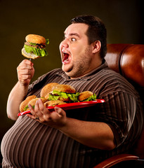 Diet failure of fat man eating fast food hamberger. Happy smile overweight person who spoiled healthy food by eating huge hamburger on fork. He's carrying a burger tray, on a leather chair. - 166409612