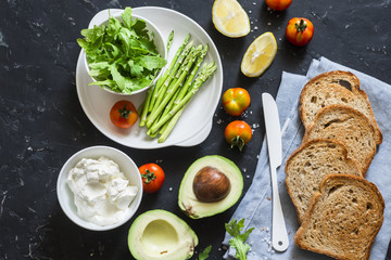 Ingredients for making toast sandwiches with avocado, asparagus, tomatoes and soft cheese on dark background, top view. Tasty breakfast, snack
