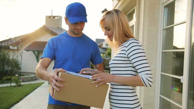 Portrait of caucasian young woman signing the documents for getting package from the courier, turning to the camera and smiling.
