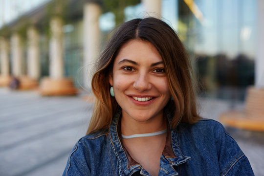 Headshot of attractive latin woman with pretty face looking and smiling cheerfully at camera, posing in urban surroundings on summer day, wearing trendy accessories and denim jacket. City lifestyle