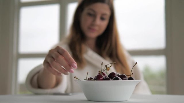 A girl in a white robe, sits on a chair, eats a cherry and uses a phone.