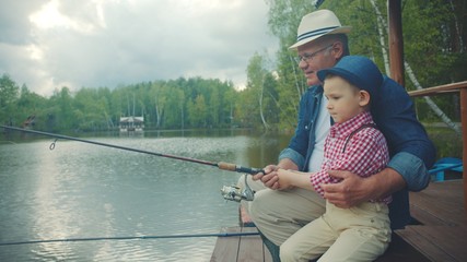 Pretty boy and his granddad are on fishing at the lake. They both wearing straw hats