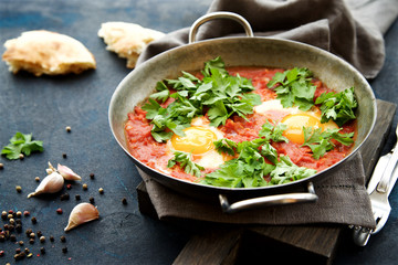 Shakshuka fried eggs with tomatoes, onions and spices in frying pan on a dark blue cracked background  
