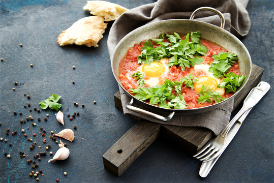 Shakshuka fried eggs with tomatoes, onions and spices in frying pan on a dark blue cracked background  
