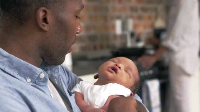 Father Holding Newborn Baby Son As Mother Prepares Meal 