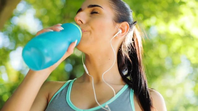 Portrait of young attractive woman with ponny tail drinking water and listening the music in the earphone, looking to the camera and smiling.