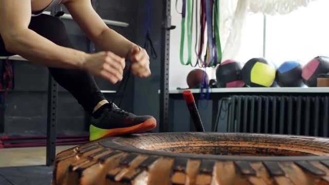 PAN with mid-section of muscular sportswoman putting her legs on tire and tying shoelaces, then working out with sledge hammer 