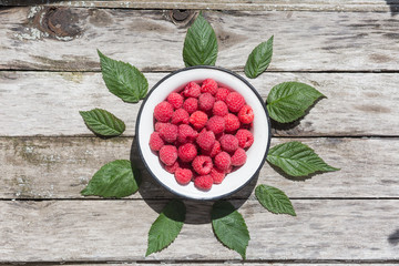 Raspberry in a white plate on an aged wood background. Green raspberry leaves. Top view.