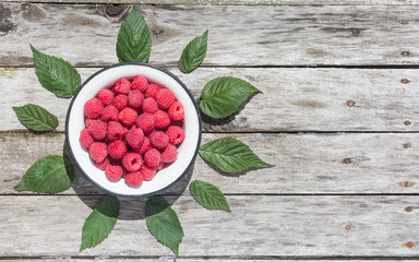 Raspberry in a white plate on an aged wood background. Green raspberry leaves. Copyspace.