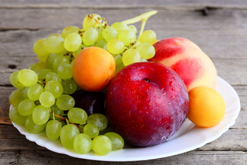 Fresh fruits assortment. Grapes bunch, peach, plum, apricot on a white plate and a wooden background. Closeup