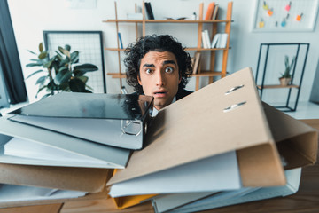 portrait of scared businessman hiding behind pile of folders while having trouble with deadline