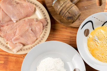 Flat lay above wooden table with raw chicken breasts mixed eggs flour and wooden hammer