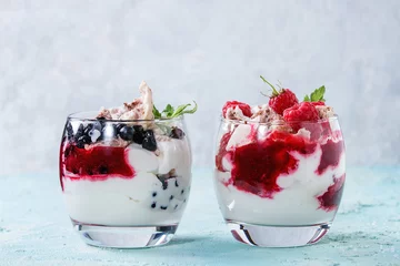 Gardinen Traditional summer dessert Eton Mess. Broken meringue with whipped cream, berry jam, fresh blueberries and raspberries in two glasses, decorated with mint leaves over light blue concrete background. © Natasha Breen