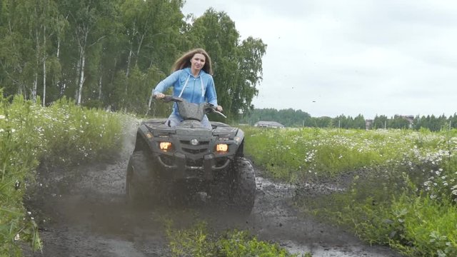 Beautiful woman rides on a quad bike along a rural road, mud is flying under the wheels, splashes