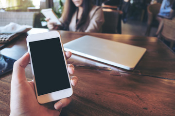 Mockup image of a man's hand holding white mobile phone with blank black screen in vintage cafe with laptop on wooden table and blur asian business woman in background