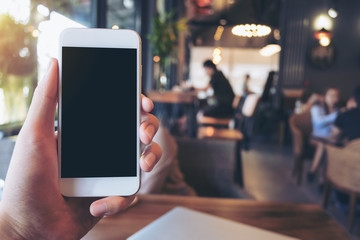 Mockup image of a man's hand holding and raising a white mobile phone with blank black screen in cafe with many people in background
