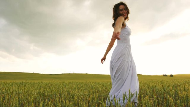 Attractive happy woman in the long white dress spinning in wheat field and smiling on camera.