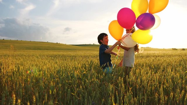Little cute boy running in the wheat field at blue sky background with balloons in the hand to his beatiful sister.