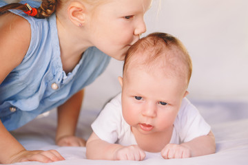 Lifestyle closeup portrait of cute white Caucasian girl sister kissing little baby, lying on bed indoors. Older sibling with younger brother newborn. Family love bonding together concept.