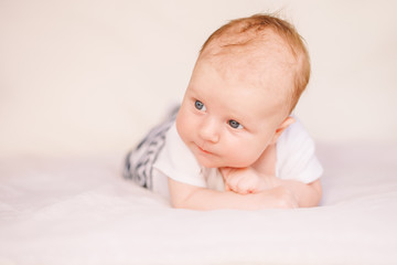 Closeup portrait of adorable funny white Caucasian baby with blue grey eyes lying on tummy belly on bed. Aware cute newborn on white light background in studio.