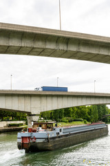 An empty cargo barge going down the river Marne toward Paris while a semi-trailer truck drives on a ramp of the A86 highway over the waterway.