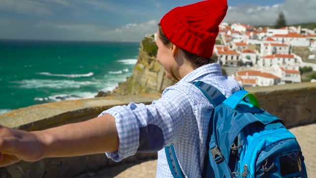 Follow me - happy young woman in a red hat and with a backpack behind her back pulling guy's hand at Azenhas do Mar, Portugal. Hand in hand walking to the ocean coast
