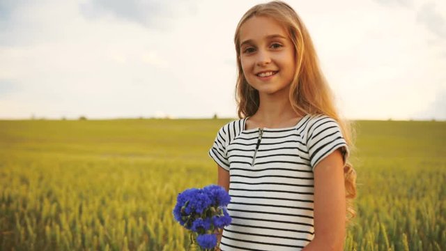 Side portrait of attractive little girl stending in the golden field with beatiful bouquet of cornflowers, turning to the camera and smiling on the camera.