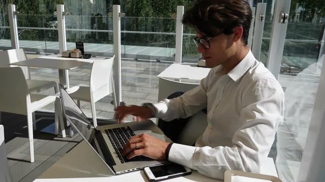 Handsome trendy young business man wearing white shirt working on laptop computer at bar table, outdoor in city setting