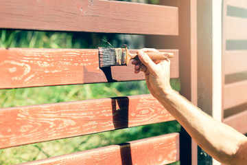 hand with paintbrush painting wooden fence with brown paint