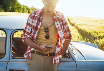 Young Man in 1970s Clothes Leaning Against Blue Vintage Car and Typing on Cellphone