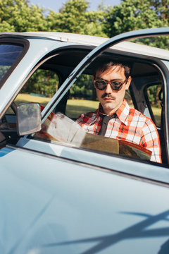 Portrait of Young Man Sitting in Blue Vintage Car and Studying Road Map on Sunny Day