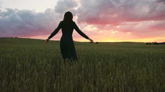 Young beatiful woman in black dress walking in the green field on the colorful sky background during the sunset.