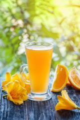 Freshly squeezed orange juice in a glass glass with orange slices and flowers yellow lilies on green bokeh background at Sunny .