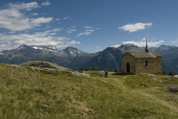 Fototapeta na wymiar Mountain summer landscape: trail with two electric bicycles E-bike on a sunny day with clouds and a small church in a high mountain village, Belalp, Alps, Switzerland
