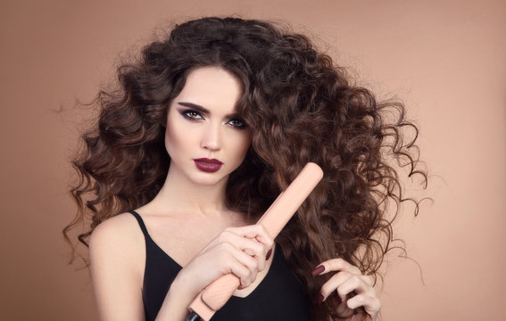 Hairstyling. Curly Beauty hair. Glamour portrait of beautiful woman model holding straightening iron. Brunette with marsala matte lips and long hairstyle isolated on beige background